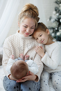 快乐的女人, 欢快的母亲和小孩, 一个女孩和一个新生的男孩坐在一个蓬松的白色毯子旁边的圣诞节日绿色的圣诞树, 白色的玩具, 雪花, 白色的蝴蝶结和发光的灯光.