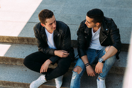 两个身穿黑色夹克、牛仔裤和白鞋的男士正坐在台阶上。在手表的手上。社交, 结交朋友.