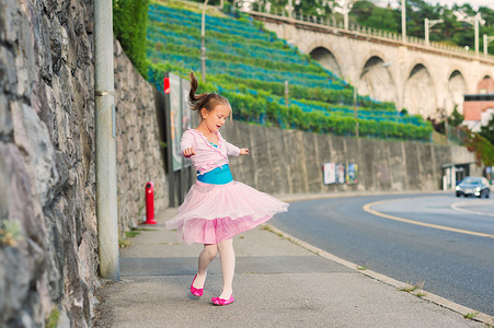 Outdoor portrait of a cute little girl of 7 years old, walking to dance school and dancing in the street, wearing purple ballet dress