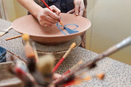 paints摄影照片_Potter woman paints a ceramic plate. Girl draws with a brush on earthenware. Process of creating clay products