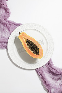 有创意的摄影照片_有创意的食物概念，半木瓜放在盘子里，紫色纺织品放在白色背景上。平坦的地方俯瞰景色.简约多彩的自然背景.