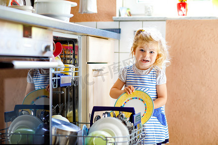 厨房碗碟摄影照片_可爱可爱的小女孩帮助卸下洗碗机。有趣而快乐的孩子站在厨房里，端着碗碟，头上放着一个碗。健康的孩子在家里。可爱的助手，玩得开心