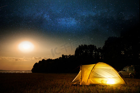 旅行和露营的概念 - 营地帐篷在夜晚的天空下满是星星。橙色照明帐篷和汽车。美丽的自然 - 田野，森林，平原。月亮和月光