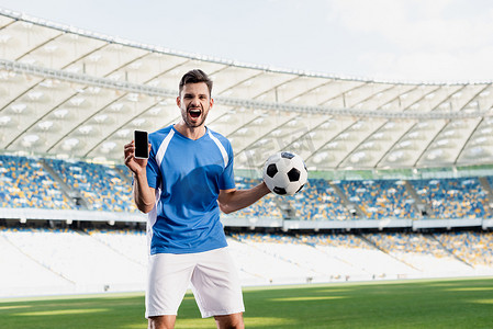 身穿蓝白相间制服的职业足球运动员，球上有智能手机，屏幕空白，在体育场大喊