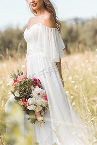 在婚礼礼服微笑的年轻新娘的被裁剪的射击在户外捧花花束