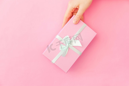 粉色礼品盒摄影照片_简单的设计女性手拿着粉红色的礼品盒隔离在粉红色柔和的色彩时尚背景