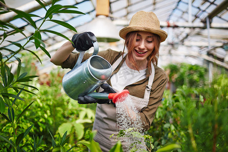 美丽的年轻女子的肖像穿着草帽享受工作在树苗圃花园, 浇水植物使用金属罐头和微笑
