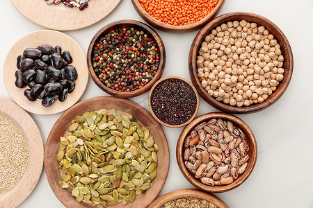 在白色大理石表面的木碗与不同的豆类，藜麦，红扁豆，胡椒粉，南瓜种子和鹰嘴豆的顶视图