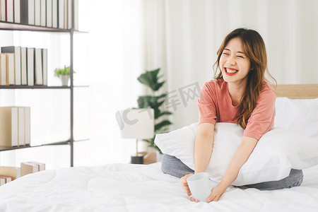 年轻的亚洲成年女子形象地坐在床上，脸上洋溢着幸福的笑容和牙齿。早上醒来手拿着咖啡杯在卧室里放松一下.