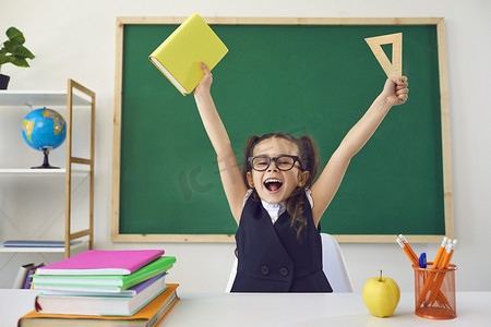 育儿培训摄影照片_回学校去一个快乐的小女生在教室的一块绿色黑板的后面举手表决.