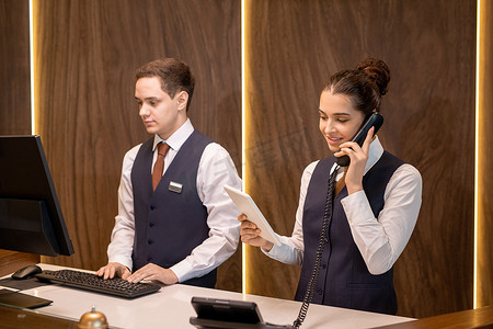 两个在柜台摄影照片_两个年轻的酒店接待员中的一个站在柜台旁，看着触摸板展览，并与同事在电话上咨询客户意见