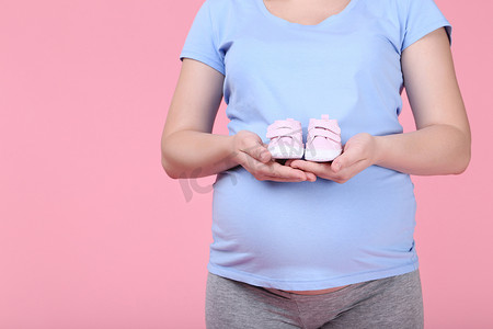 粉红色孕妇摄影照片_在粉红色背景下抱着婴儿鞋的孕妇