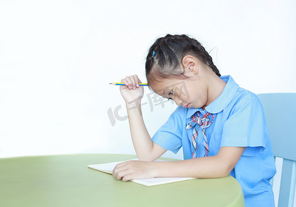 紧张的亚洲小女孩学习，厌倦了在白底的桌子上做作业。 小女孩用铅笔捂住头.