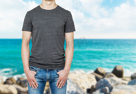 shirt摄影照片_Close-up of a man in a blank grey t-shirt. Hands in the denims pockets. Blur sea view.