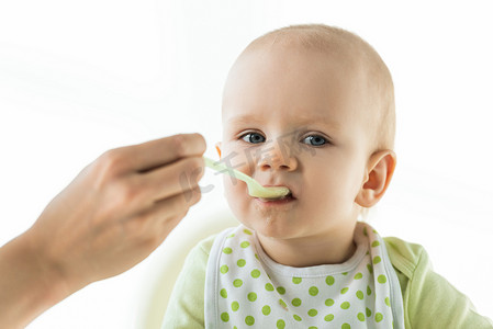 Selective focus of mother with spoon feeding baby with puree on feeding chair isolated on white