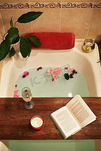 为你自己的时间。在家里放轻松用花瓣沐浴浴缸。木盘上的书,蜡烛和酒杯.舒适舒适浴室中的有机温泉放松