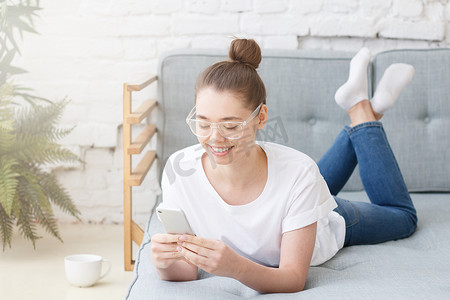 漂亮的欧洲白种女孩的水平图片躺在灰色沙发上有塑料透明眼镜手持手机, 与朋友聊天或浏览移动互联网