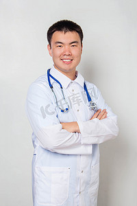 一位身穿白衣、脖子上戴听诊器的年轻的亚洲医生站在那里，双手插在胸前，背景是白色的.