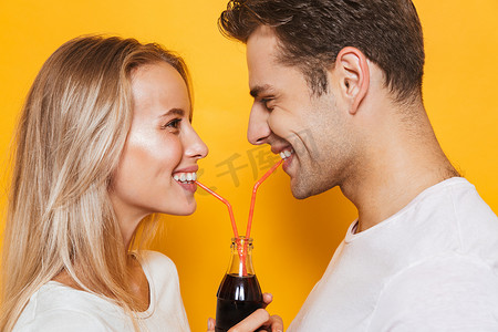 漂亮的年轻夫妇站在黄色的背景下, 喝碳酸饮料从玻璃瓶, 看着对方