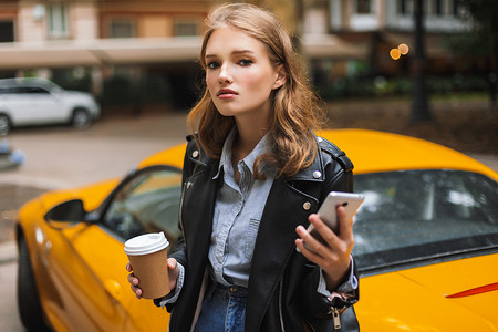 年轻有吸引力的妇女在皮革夹克拿着杯咖啡去和手机在手朦胧地看着黄色跑车在城市街道背景的相机