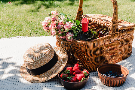 白毯子摄影照片_在阳光明媚的日子里，在花园中，在草帽和浆果附近的白毯子上，用玫瑰和一瓶葡萄酒装着柳条篮
