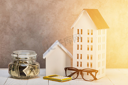 房子模型在白色木桌上与眼镜,计算器和钱箱在阳光下,房地产概念