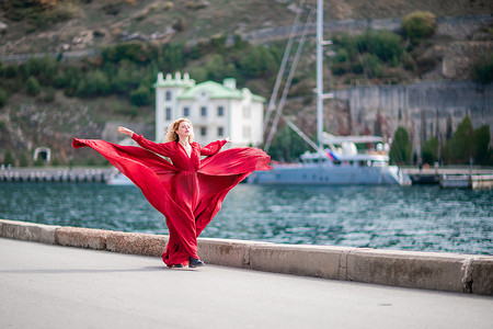 一个穿着红色连衣裙的女人，一个穿着飘逸的长丝翅膀的时装模特，在堤岸上飘扬着面料.