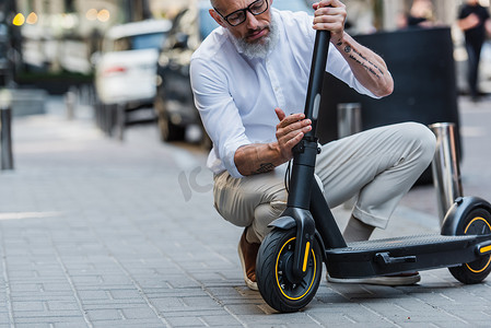 mature man in glasses and shirt adjusting electric scooter on street 