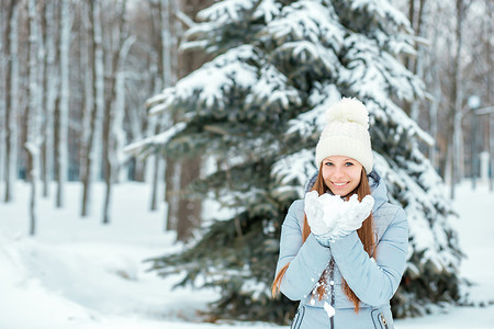 帽子冬天摄影照片_一个女孩穿着暖和的冬天的衣服和帽子在冬天的森林里吹雪, 水平。模型与美丽的微笑在圣诞树附近.