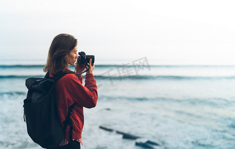 海洋模糊摄影照片_带背包的时髦徒步旅行者用相机拍摄的令人惊叹的海景日落在背景蓝海, 摄影师享受海洋地平线, 模糊的全景日出毛坯样机, 旅客放松假期概念, 在旅游度假的阳光景观 