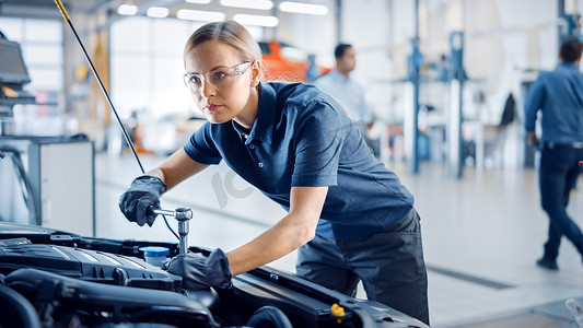 美丽的增强女性力量的机修工是在汽车服务公司的汽车上工作。戴安全眼镜的女人正在修理发动机.她用的是斧头。现代汽车清洁工场.