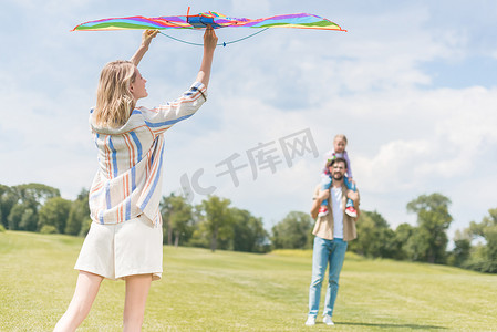 五颜六色的风筝摄影照片_妇女抱着五颜六色的风筝, 而父亲背着小女儿在脖子后面的公园 