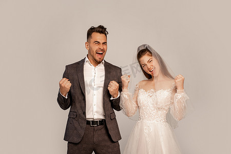win摄影照片_excited newlyweds showing win gesture while looking at camera isolated on grey