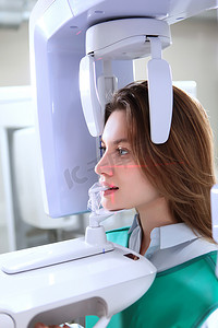 背心摄影照片_穿着防护背心的病人用全景X光检查牙齿。医生把检查牙齿的设备开了一半.诊所里的X光设备现代医学。全景X光的牙齿。侧视图.垂直照片.