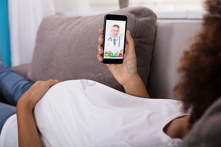 怀孕的妇女摄影照片_怀孕的妇女躺在沙发上视频会议与男性医生的智能手机
