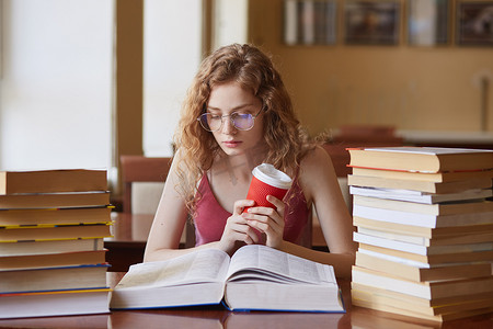 大学生支教摄影照片_教育和学校理念。聪明的女学生手拿咖啡，戴着眼镜，穿着休闲装，神情专注，坐在大学图书馆的书桌上，拿着书堆看书.