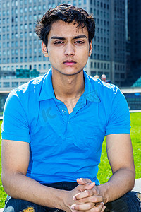 短袖蓝色摄影照片_18岁的美国东印度群岛青少年在纽约市的肖像。年轻的男大学生穿着蓝色短袖衬衫，坐在商业区的绿色草坪旁，放松地思考着
