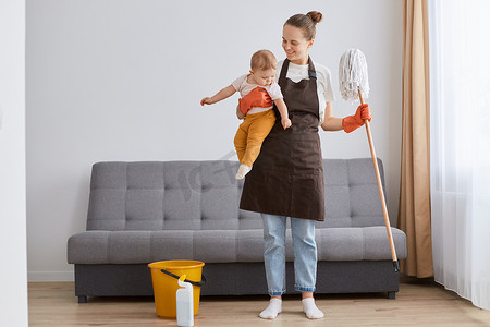 Portrait of smiling positive woman wearing brown apron with mop and little toddler daughter in hands cleaning in living room, housewife in maternity leave..