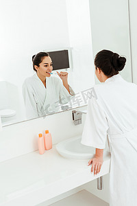刷牙摄影照片_选择焦点的快乐年轻女子刷牙，看着镜子在浴室 