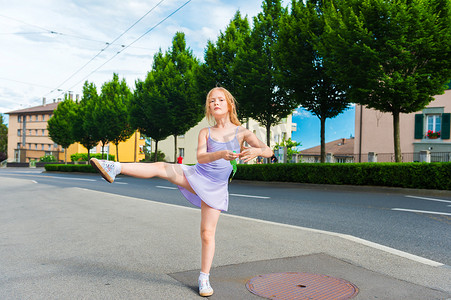 dance摄影照片_Outdoor portrait of a cute little girl of 7 years old, walking to dance school and dancing in the street, wearing purple ballet dress