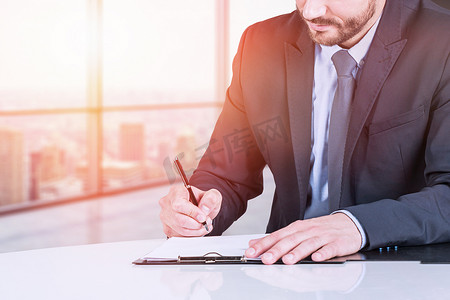Unrecognizable bearded businessman writing in clipboard in panoramic office with cityscape. Paperwork concept. Toned image