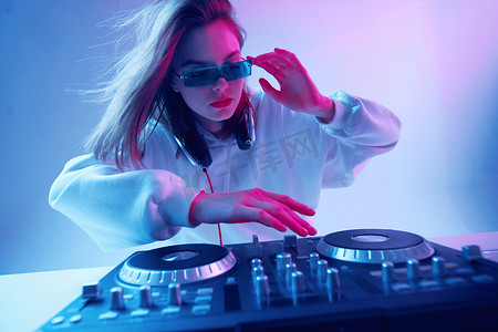 酷酷眼镜摄影照片_年轻酷女孩DJ在混合控制台和耳机上混合着音乐，穿着时髦的衣服，戴着霓虹灯背景的眼镜.