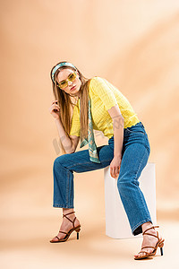 穿着牛仔裤、黄色T恤、太阳镜、高跟鞋和丝巾的漂亮女孩坐在米黄色的白色立方体上