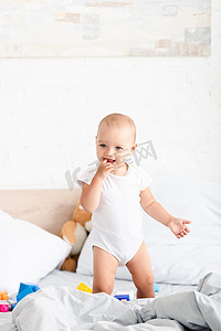 可爱的赤脚婴儿在白色的衣服站在床上与玩具，并采取手到嘴