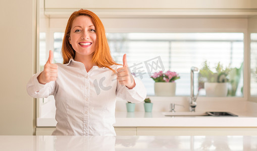 大拇指向上摄影照片_红头发的女人在厨房成功的标志做积极的手势与手, 大拇指向上微笑和快乐。看着带着欢快表情的相机, 胜利者的手势.