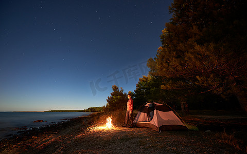 在海边过夜露营。徒步旅行的妇女休息在篝火旁的旅游帐篷和森林在星空下, 享受清澈的蓝色水的美丽景色。旅游与积极的生活方式理念.