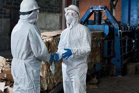 两名身穿生物防护服的工人肖像在现代废物处理厂工业仓库中对可回收纸板进行通信在后台, 复制空间