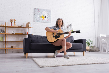 一个快乐而成熟的女人，灰白的头发坐在沙发上，在客厅里弹奏着吉他