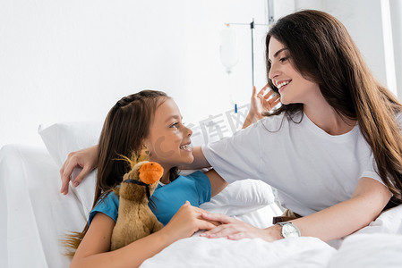 积极的孩子抱着妈妈，在诊所的床上抱着柔软的玩具 