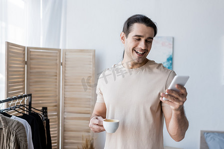 T恤摄影照片_穿着T恤的快乐男人拿着一杯咖啡，用智能手机